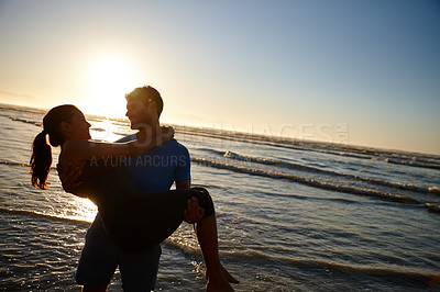 Buy stock photo Shot of a young man carrying his girlfriend on the beach at sunrise
