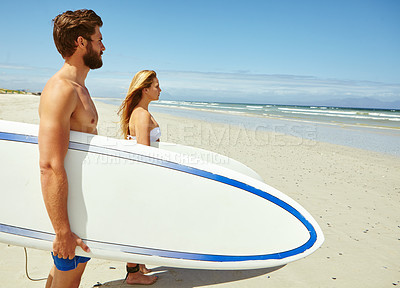 Buy stock photo Shot of a young couple standing on the beach with their surfboards looking out at the ocean