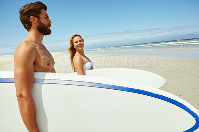 Buy stock photo Shot of a young couple walking on the beach, carrying their surfboards