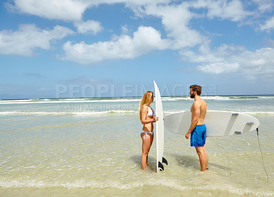 Buy stock photo Shot of a young couple standing with their surfboards in shallow water at the beach