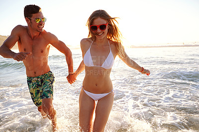 Buy stock photo Shot of a young couple in swimwear running through waves at the beach