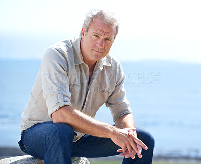 Buy stock photo Mature, man and outdoor portrait at the sea on holiday, vacation or travel in nature to relax. Serious, person and calm ocean, water or waves in morning and sitting on a rock with blue sky in summer