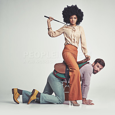 Buy stock photo A studio shot of an attractive woman in 70s wear riding a handsome man wearing a saddle while using a riding crop
