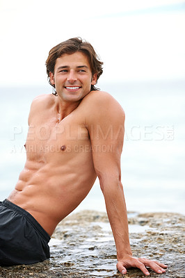 Buy stock photo Shot of a handsome young man sitting on a rock at the beach