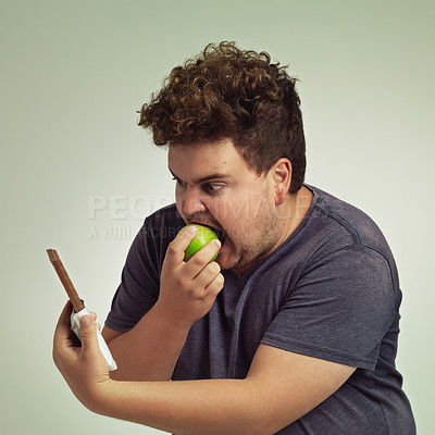 Buy stock photo Guy, apple and chocolate in studio with choice of junk food, sweet or diet for wellness. Plus size, male person eating and decision, fruit or candy for nutrition, lose weight and healthy lifestyle