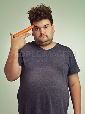 Buy stock photo Portrait, carrot and hand gun with plus size man in studio on gray background for diet, health or nutrition. Depression, sad  and vegetable to head of young person in conflict with vegetarian food