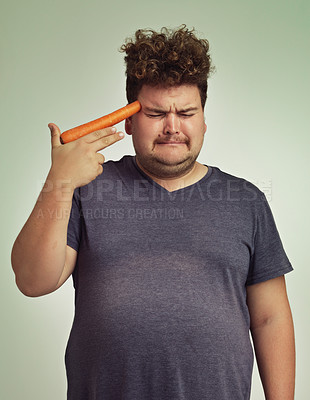Buy stock photo Depression, vegetable and hand gun with plus size man in studio on gray background for diet, health or nutrition. Sad, unhappy and carrot to head of young person in conflict with vegetarian food