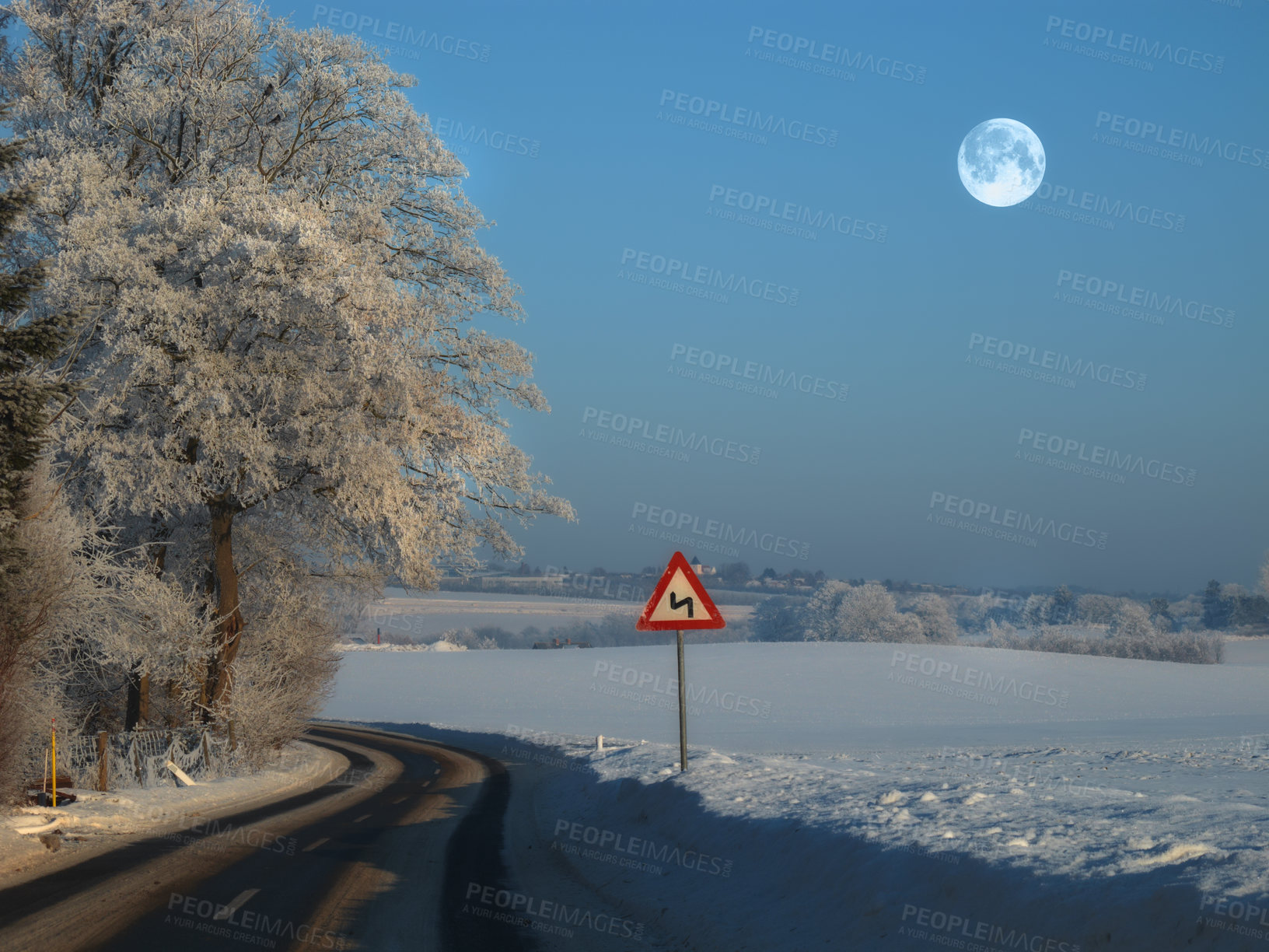 Buy stock photo The winding road on a rural winter landscape with a sign on the side of the empty road for safety. A slippery and wet road surrounded by snow on a cold winter evening near a forest with ice frost