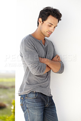 Buy stock photo Shot of a handsome young man leaning against a wall with his arms crossed