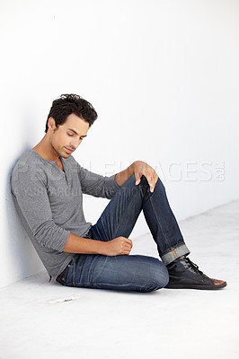 Buy stock photo Shot of a young man looking thoughtful while sitting on the floor leaning against a white wall