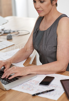 Buy stock photo Cropped image of an architect working on her laptop with building plans beside her on her desk