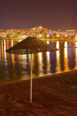 Buy stock photo Umbrella, beach and night with city lights, reflection and vacation with adventure, travel and getaway trip. Empty, evening and nature with water, sand and holiday with journey, dark and environment