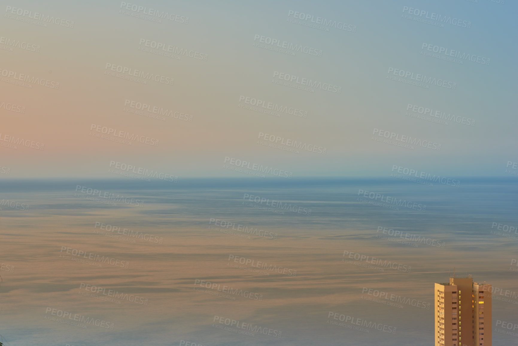 Buy stock photo Calm waters during sunset with a hotel building overlooking the ocean. Aerial view of the sea during dusk. Copy space of small waves on calm water in the sea, bright afterglow in a clear summer sky 
