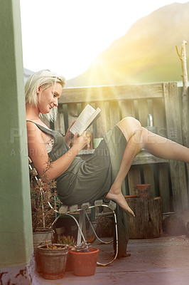 Buy stock photo Shot of a young woman reading a book while sitting on a balcony in the sunlight