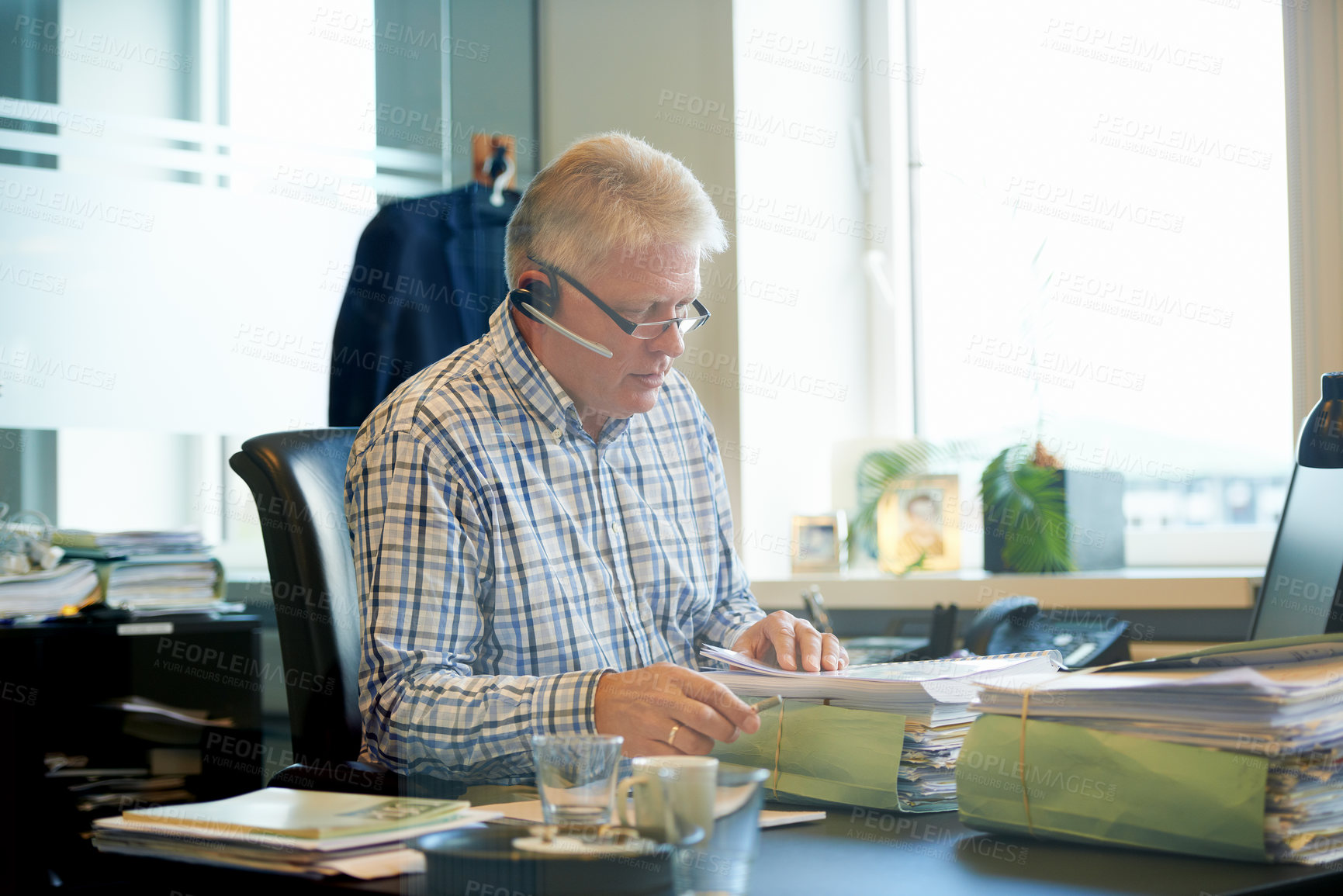 Buy stock photo Shot of a senior businessman going through paperwork at his desk while wearing a headset