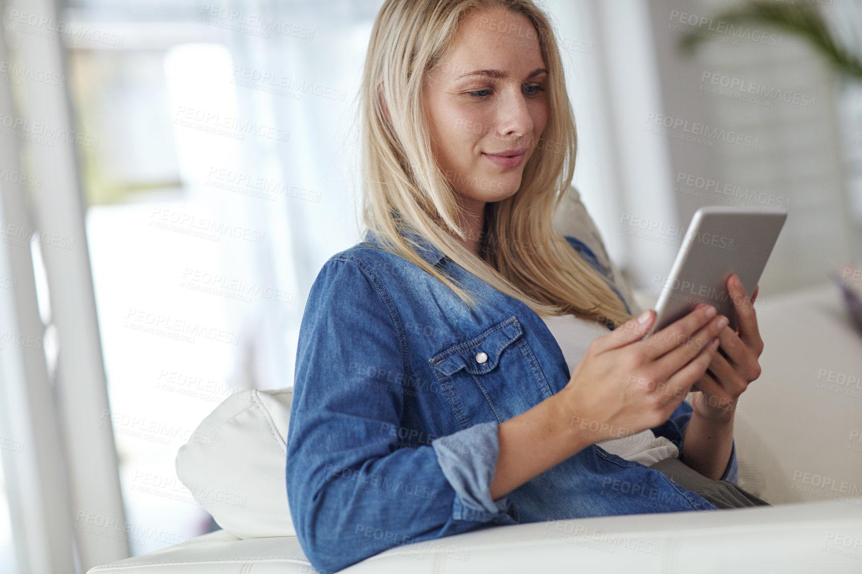 Buy stock photo Shot of a young woman using a digital tablet while sitting on her living room sofa