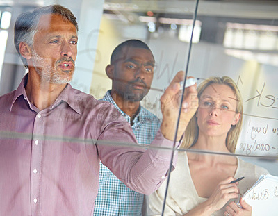 Buy stock photo A mature businessman writing down plans on a glass pane while his colleagues look on
