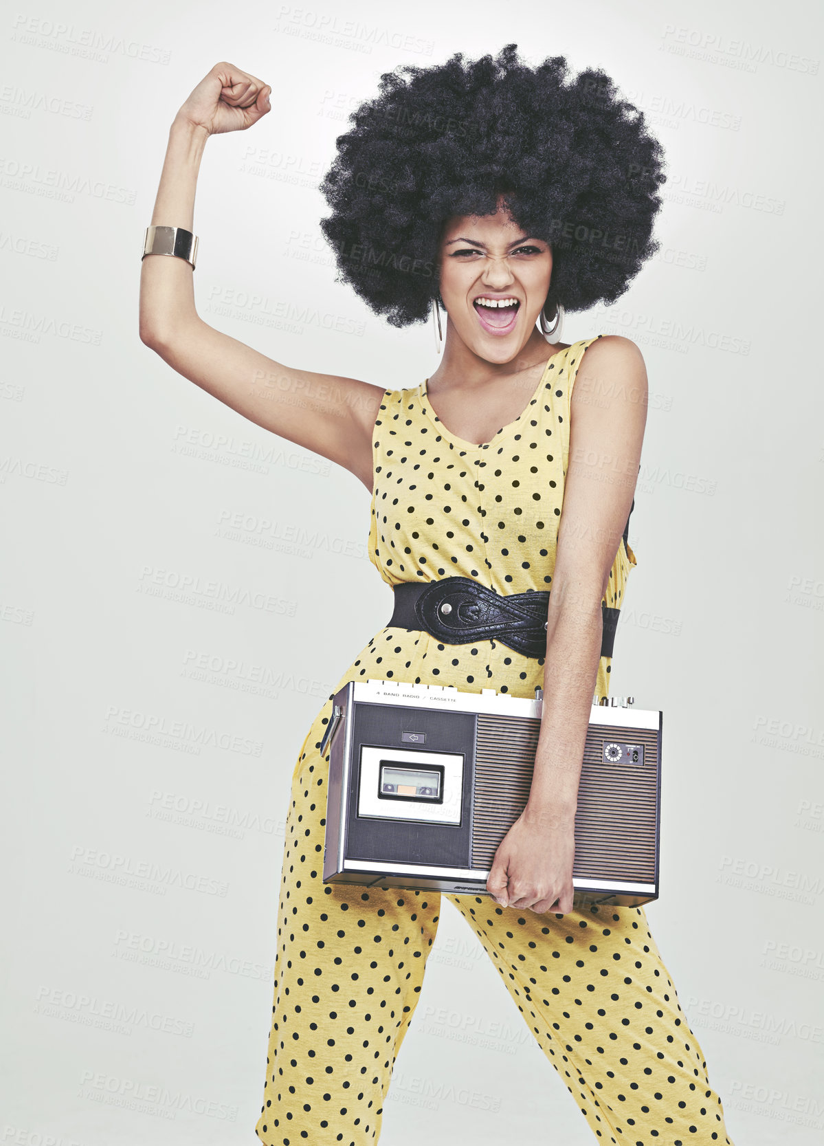 Buy stock photo A young woman wearing a 70s retro jumpsuit holding a cassette player and striking a disco pose