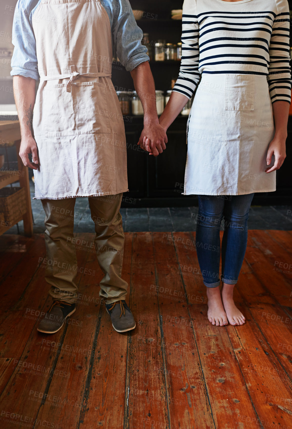 Buy stock photo Home, couple and holding hands with apron in kitchen for cooking support, love and bonding with baking. People, feet and culinary relationship in house for commitment, loyalty and trust with floor