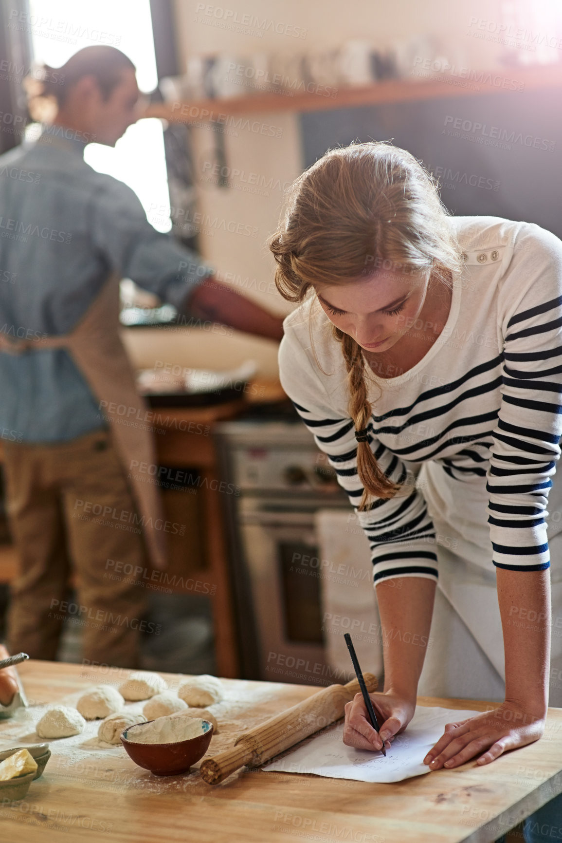 Buy stock photo Cropped shot of a woman writing a recipe for the dough that she's preparing with her boyfriend in the background