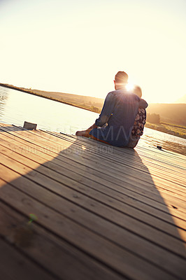 Buy stock photo Rearview shot of an affectionate young couple sitting on a dock at sunset