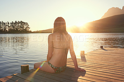 Buy stock photo Shot of a young woman in a bikini sitting on a dock by a lake at sunset