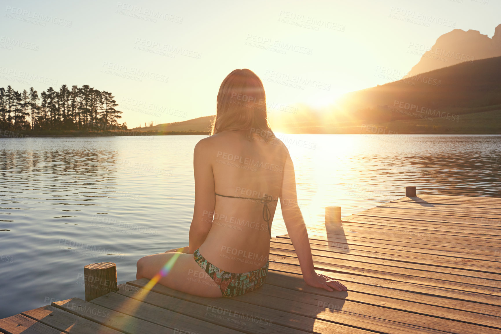 Buy stock photo Shot of a young woman in a bikini sitting on a dock by a lake at sunset