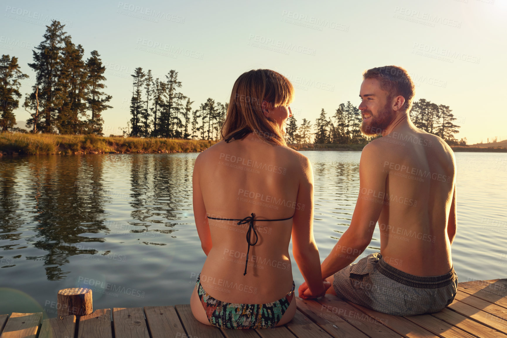 Buy stock photo Couple, holding hands and sunset on pier by river for memory, vacation or love in countryside. People, man and woman with back, water and view in nature with bikini, relationship or holiday in Italy