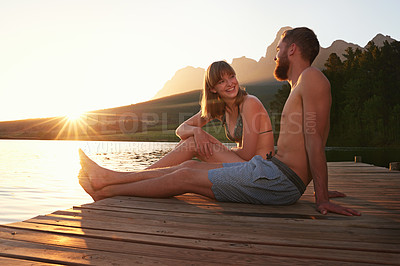 Buy stock photo Shot of an affectionate young couple in swimsuits sitting on a dock at sunset