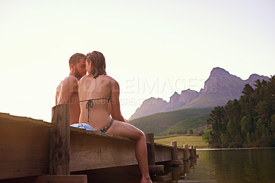 Buy stock photo Rearview shot of an affectionate young couple in swimsuits sitting on a dock at sunset