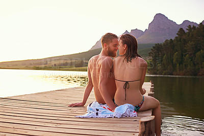 Buy stock photo Rearview shot of an affectionate young couple in swimsuits sitting on a dock at sunset