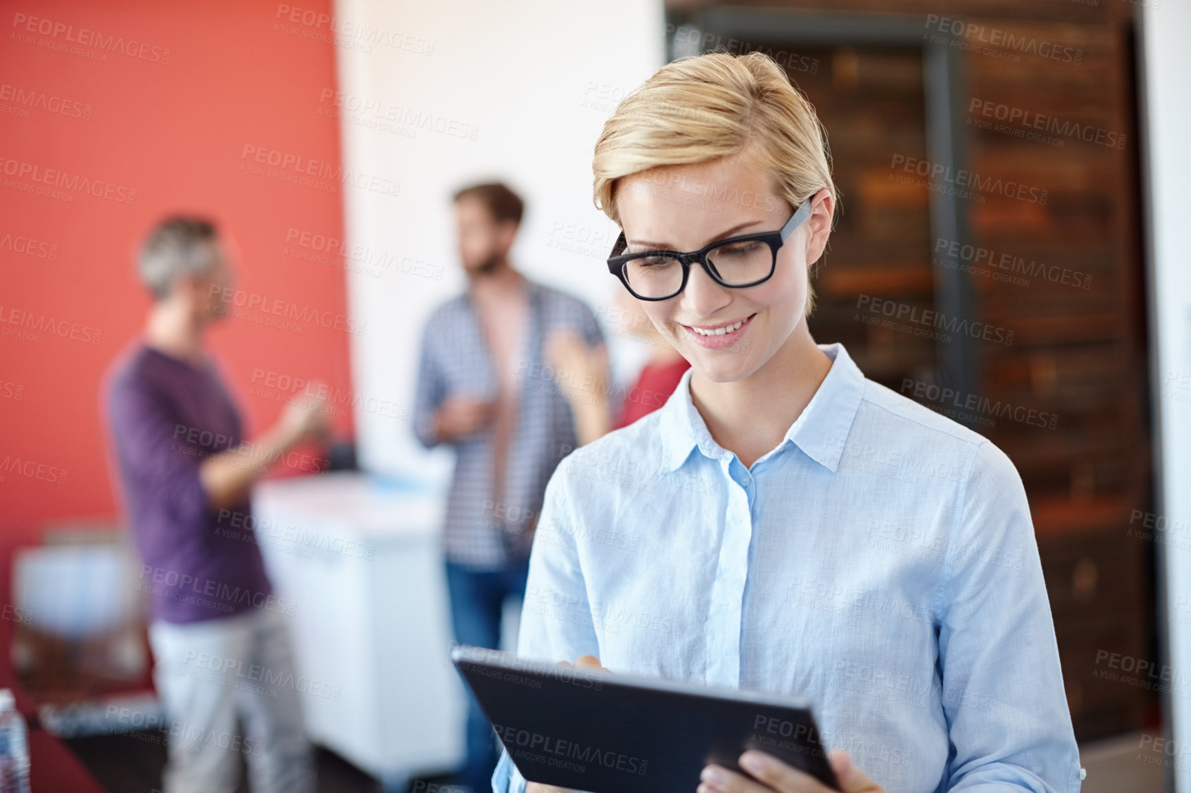 Buy stock photo Shot of a young designer using a tablet with her colleagues standing in the background