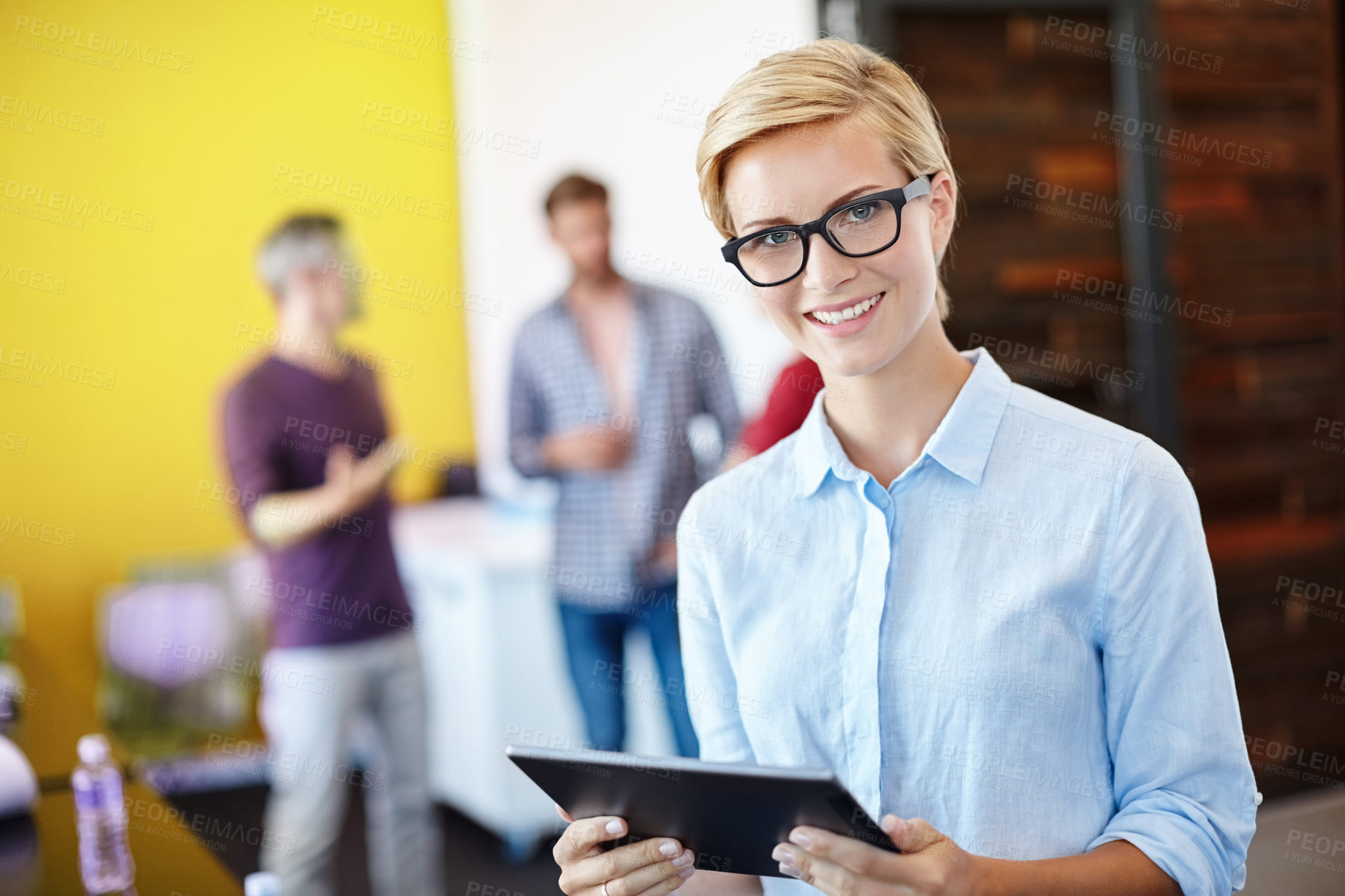 Buy stock photo Shot of a young designer using a tablet with her colleagues standing in the background