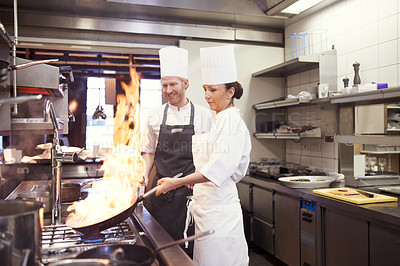 Buy stock photo Chef, people and fire with frying pan in kitchen, catering service and prepare food for fine dining. Professional, cooking and man and woman flambe and smile for teamwork in commercial restaurant