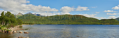 Buy stock photo Landscape of a lake with trees near a mountain. Green hills by the seaside with a blue sky in Norway. A calm sea near a vibrant wilderness against a bright cloudy horizon. Peaceful wild nature scene