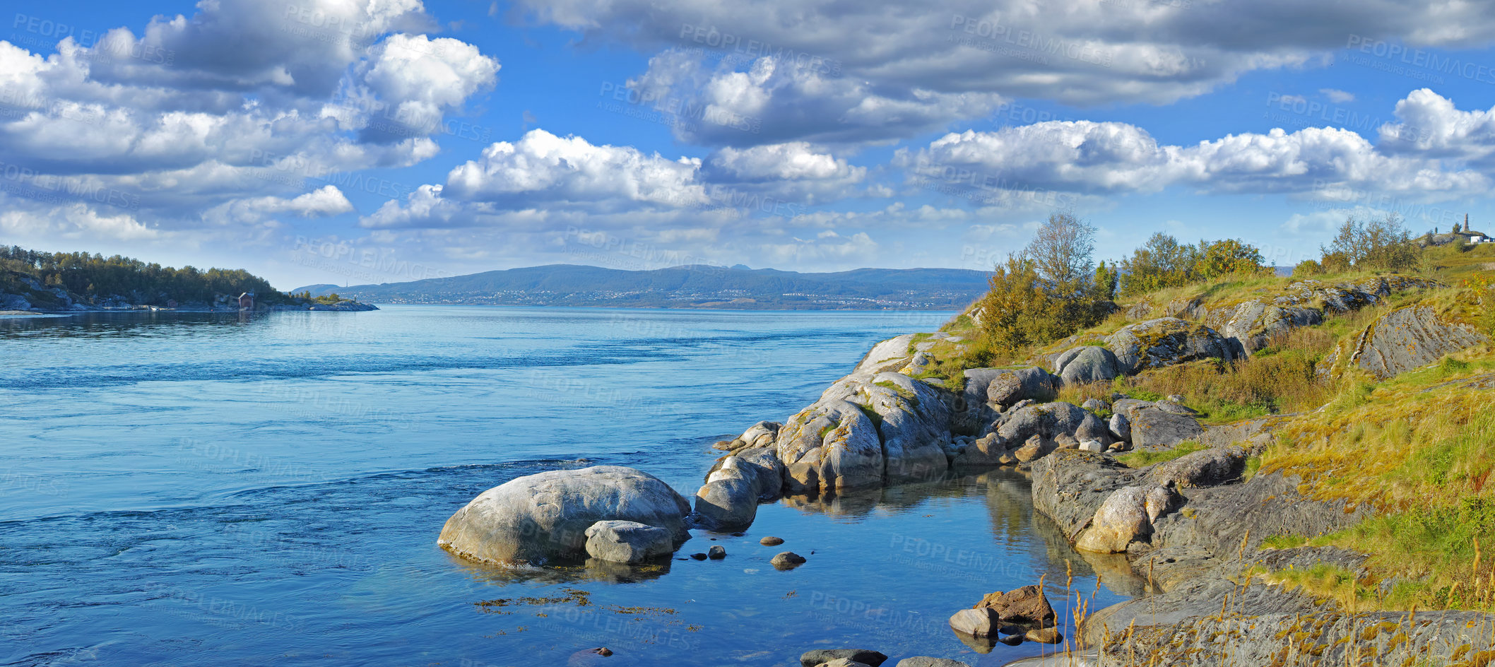 Buy stock photo Landscape of a lake with a rocky shore on a bright cloudy day. Green hill with boulders by the seaside with a blue sky in Norway. A calm serene horizon of a vibrant peaceful nature scene in Nordland