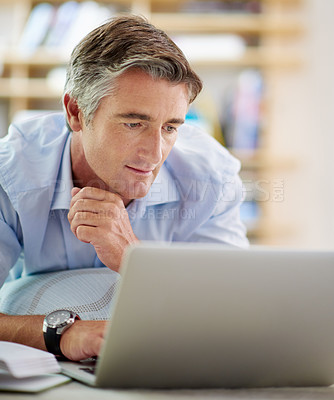 Buy stock photo Shot of a handsome mature man lying on his living room floor using a laptop