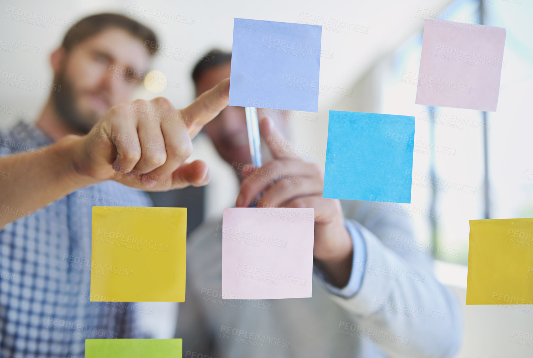 Buy stock photo Through the glass shot of two male coworkers arranging sticky notes on a wall during a meeting