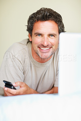 Buy stock photo Portrait, phone and laptop with a man in the bedroom of his home to relax on weekend time off. Smile, tech and a man lying in bed, streaming a movie or series for entertainment while mobile browsing 