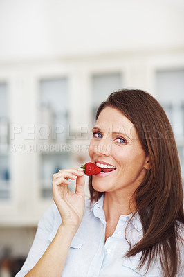 Buy stock photo An attractive brunette eating a strawberry while standing in a kitchen