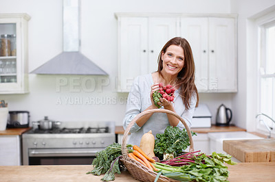 Buy stock photo A gorgeous woman holding up a bunch of radishes while standing behind a basket of fresh vegetables in her kitchen