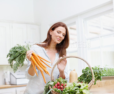 Buy stock photo A gorgeous woman holds a bunch of carrots while looking into her basket of vegetables in her kitchen