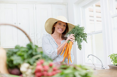 Buy stock photo A beautiful woman with a hat holding a bunch of carrots in her kitchen with a basket of vegetables in the foreground