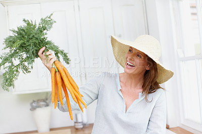Buy stock photo A beautiful woman wearing a hat holds up a bunch of carrots  in her kitchen