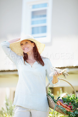 Buy stock photo A beautiful woman holds her gardening gloves and a basket of freshly picked vegetables while standing in her garden
