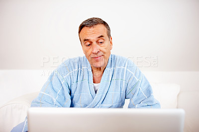 Buy stock photo Gown, morning and mature man on laptop reading email, social media or streaming subscription on sofa. Internet search, couch and senior person in bathrobe on computer, website or networking in home.