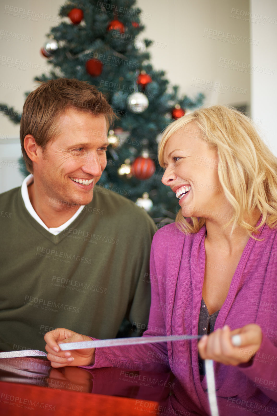 Buy stock photo Happy couple, festive season and ribbon for gift wrapping by christmas tree, love and care in house. Woman, man and smile for presents for together for giving, kindness and bonding in home on holiday