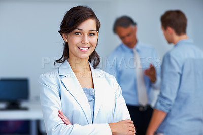 Buy stock photo Portrait of confident young business woman standing with arms crossed with staff discussing in background