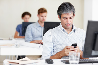 Buy stock photo Shot of an office worker sending a text message while sitting at his workstation with coworkers behind him