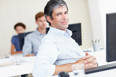Buy stock photo Portrait of a confident mature office worker sitting at his workstation with colleagues sitting behind him